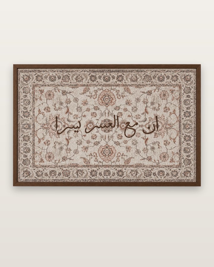 Inna ma’al usri yusra in brown Arabic calligraphy on a light beige floral canvas, in a walnut-tone frame